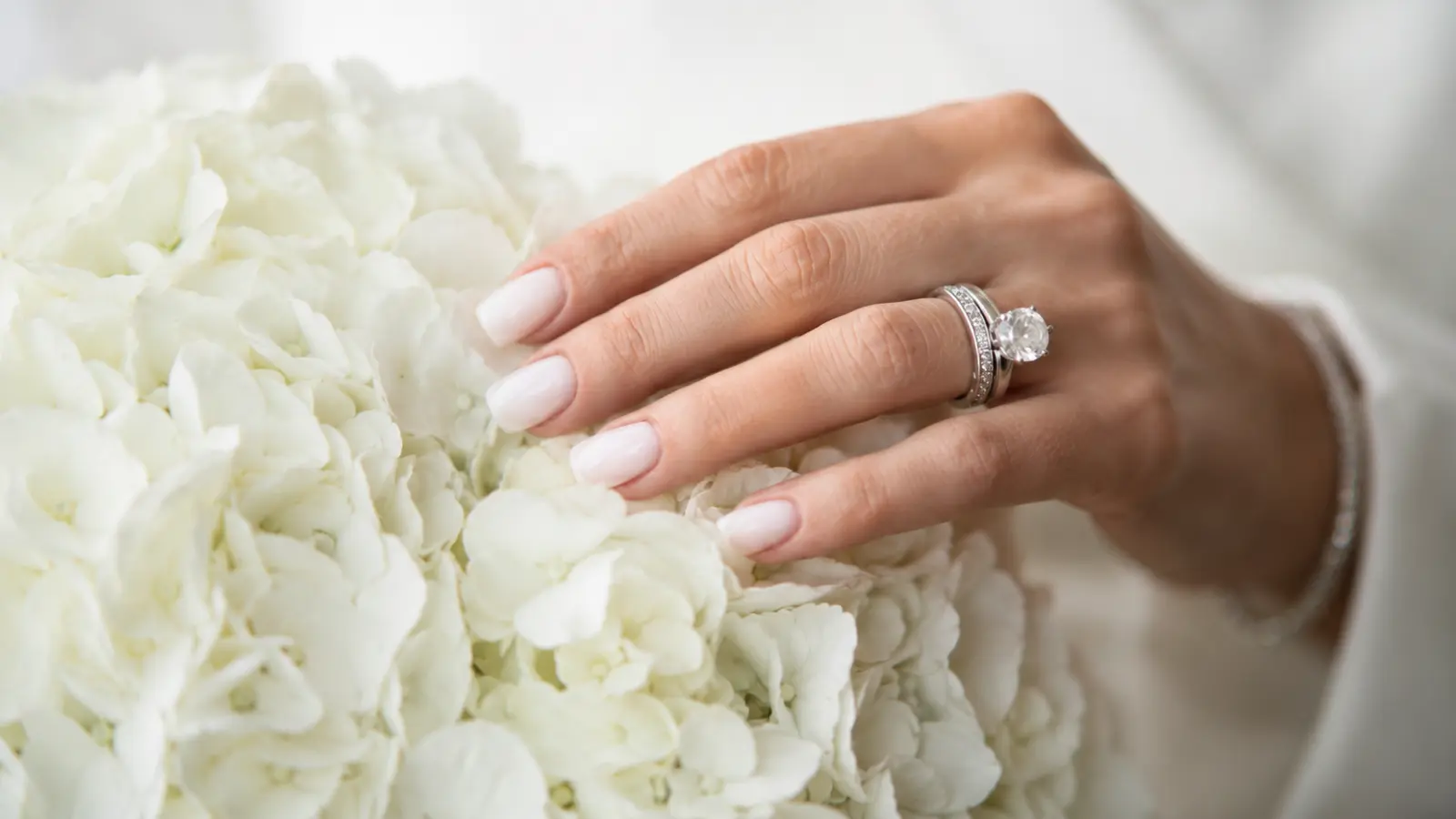 Soft white wedding nails resting on white bridal flowers with diamond ring