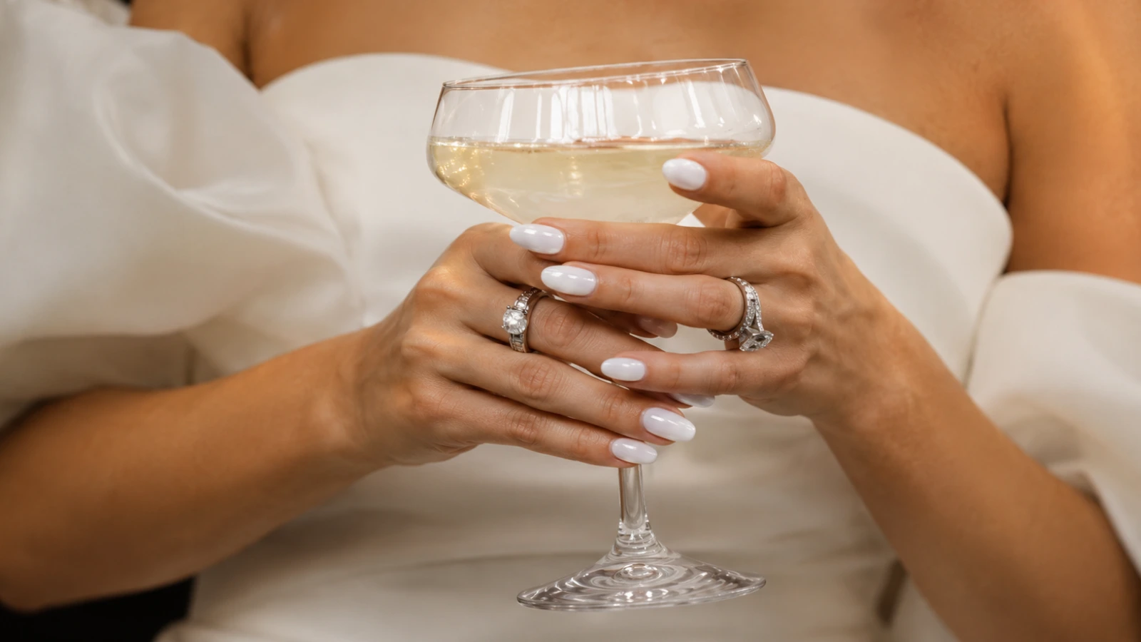 Bride holding a champagne glass with elegant white wedding nails and diamond rings
