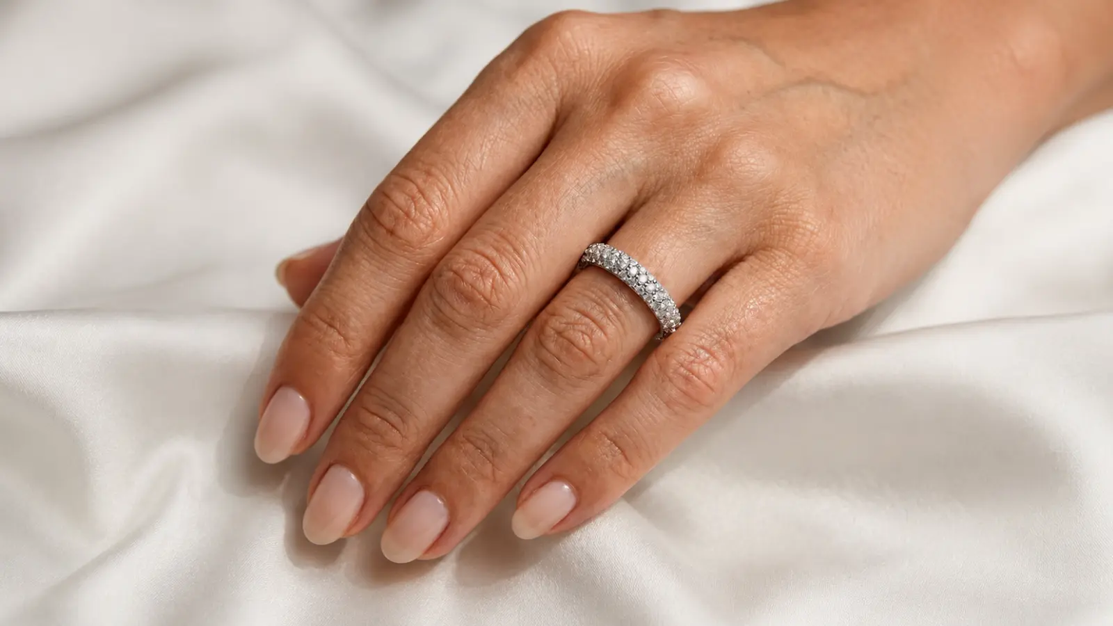Close-up of a woman's hand wearing a white gold diamond eternity wedding band on white satin fabric