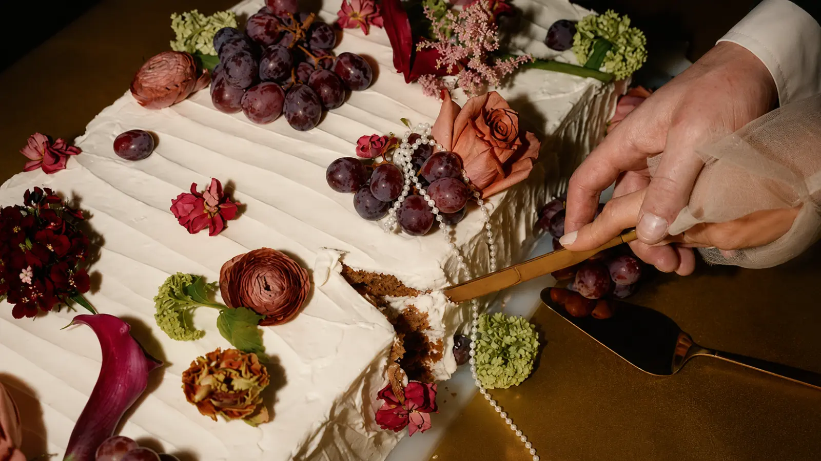 Fall wedding cake being cut with a gold knife, decorated with grapes, autumn flowers, and romantic seasonal details for an elegant wedding reception.