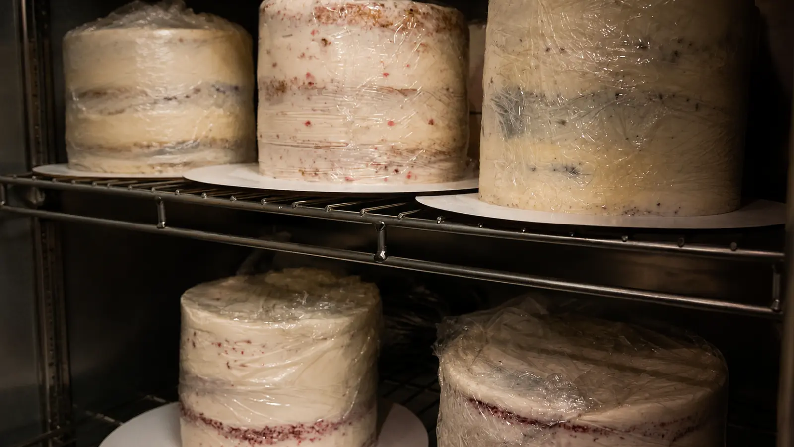 Multiple wedding cake tiers wrapped in protective plastic and stored in a freezer to preserve the cake layers after the wedding day.