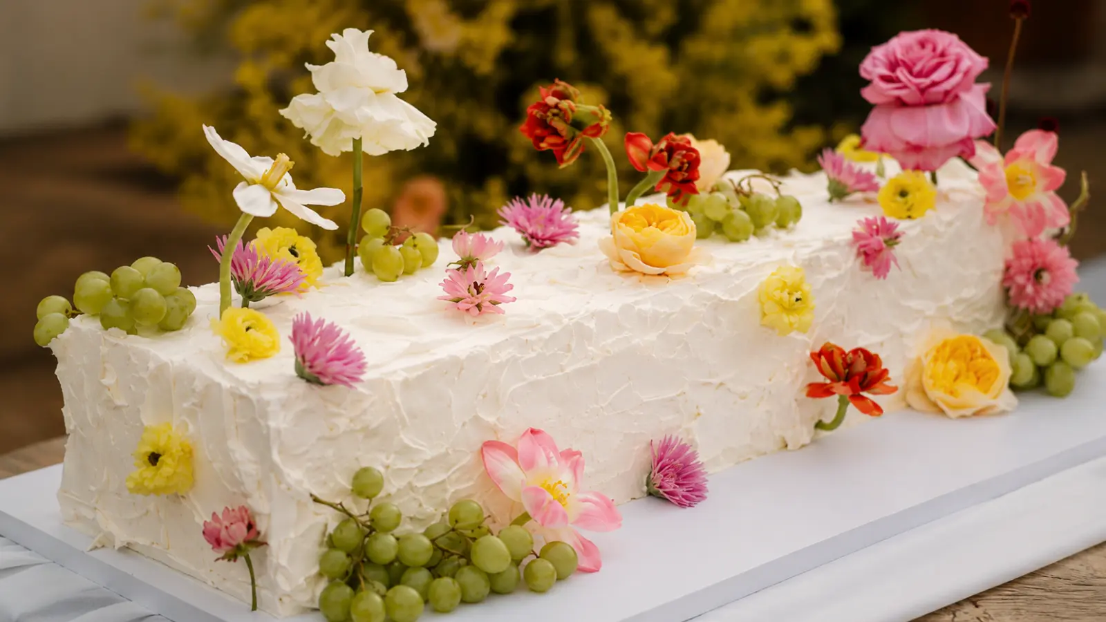 Long Italian wedding cake with textured white frosting, colorful fresh flowers, and grapes arranged in an elegant garden-style wedding design.