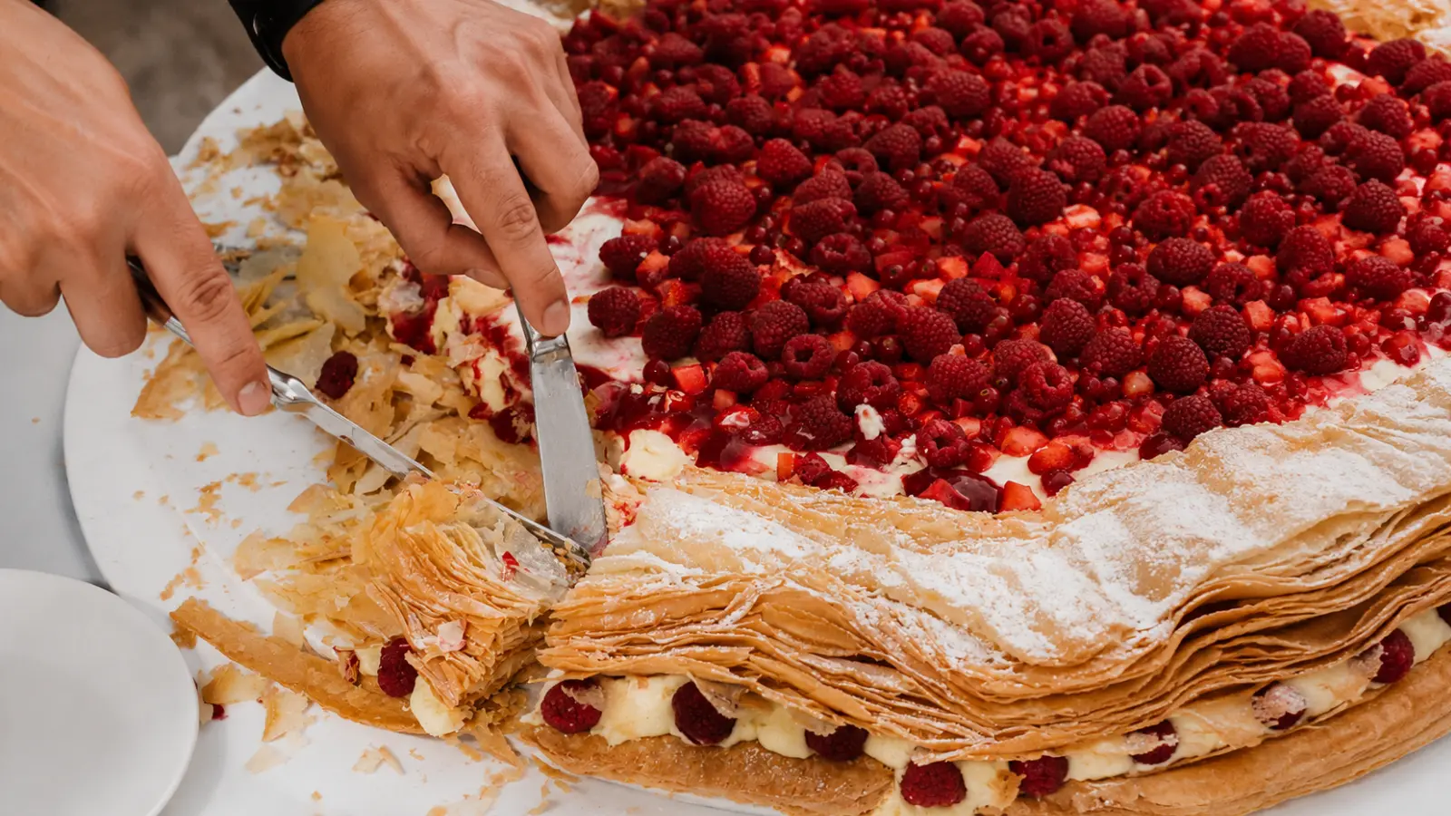 Close-up of a traditional millefoglie italian wedding cake being cut, showing crispy pastry layers filled with cream and topped with fresh raspberries and berries.