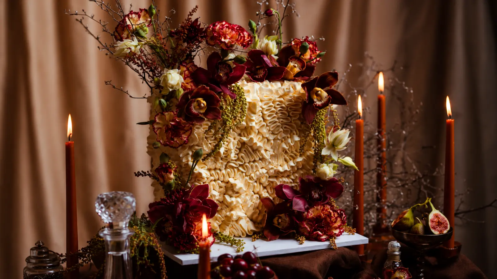 Moody fall wedding cake with textured frosting, burgundy floral arrangements, candles, and warm autumn colors for an elegant seasonal wedding display.