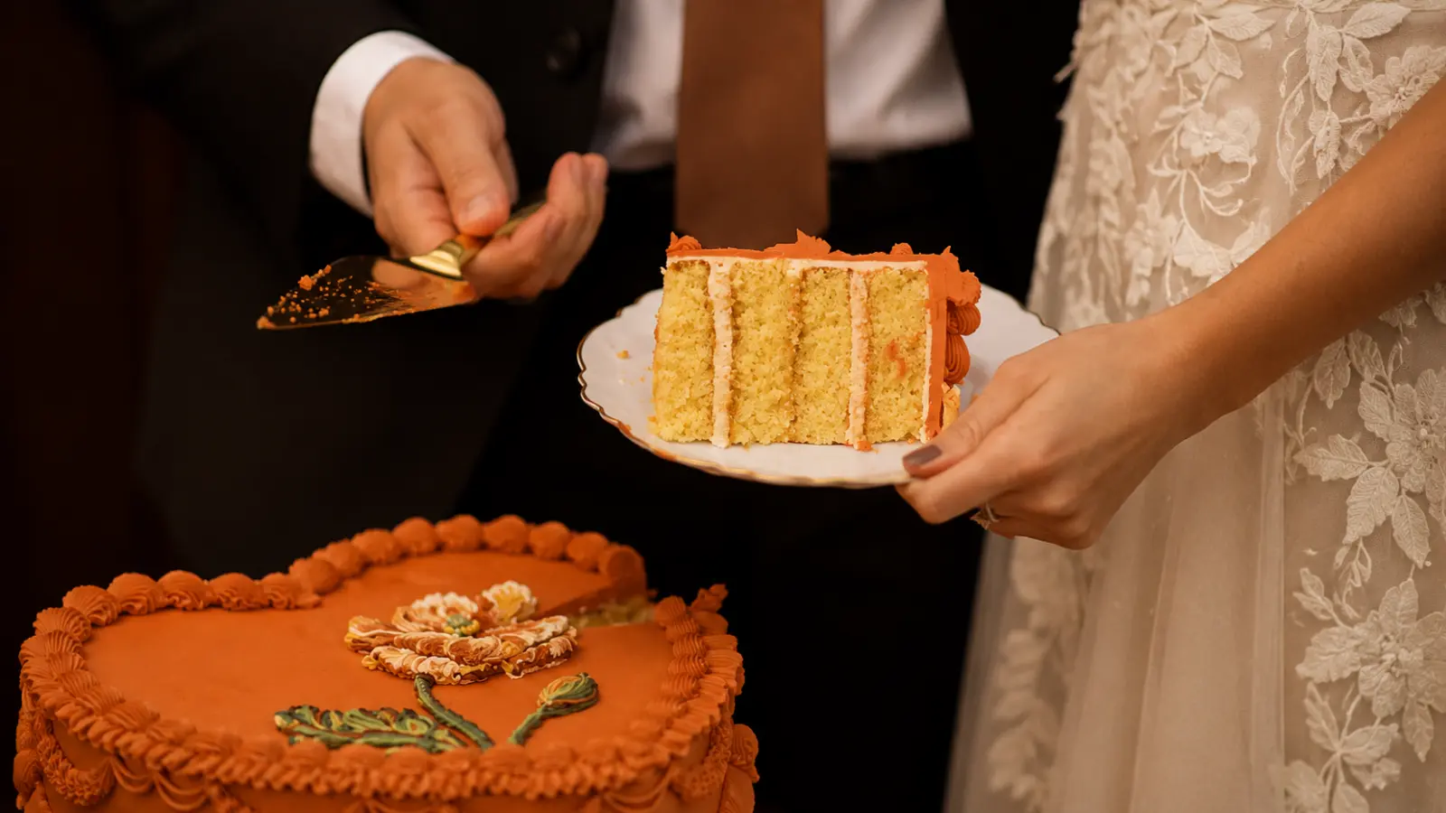 Orange fall wedding cakes with vintage piping details and floral decoration while a slice is served during an autumn wedding celebration.