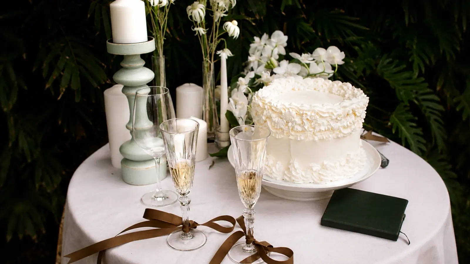 Simple white wedding cake displayed on a reception table with classic frosting details, candles, flowers, champagne glasses, and minimalist wedding decor.