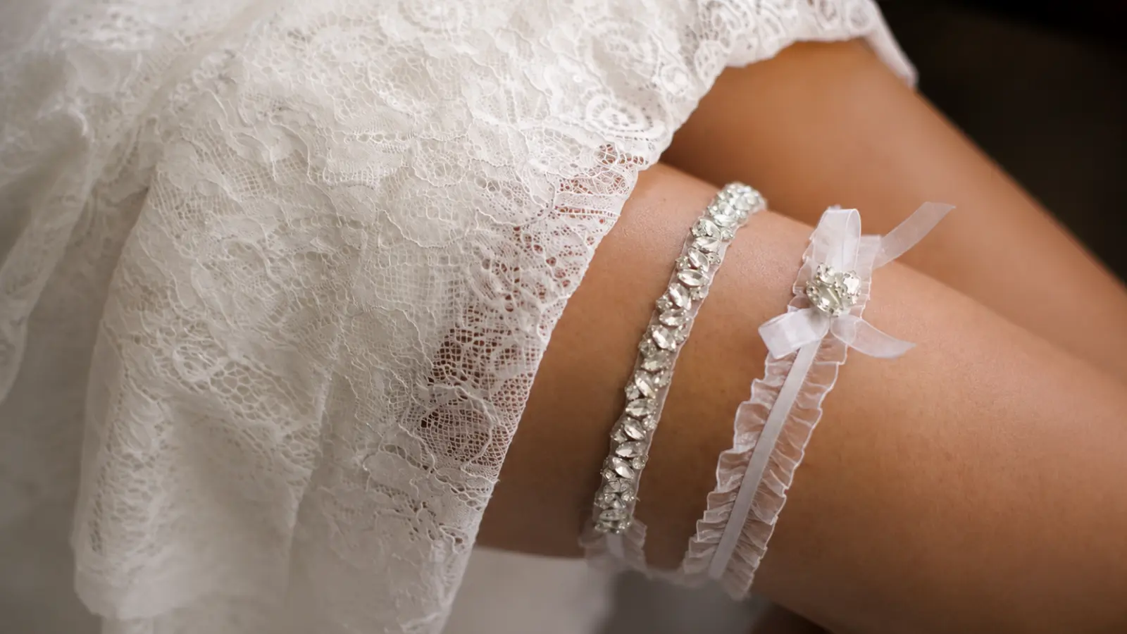Two wedding garters with different lace and crystal designs displayed beneath a lace wedding dress