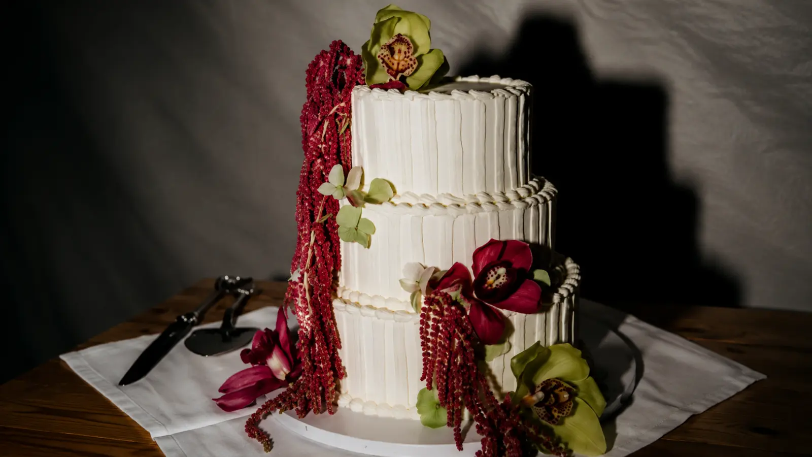 Three-tier fall wedding cake with white textured frosting, burgundy flowers, greenery, and elegant autumn decorations on a rustic table setting.