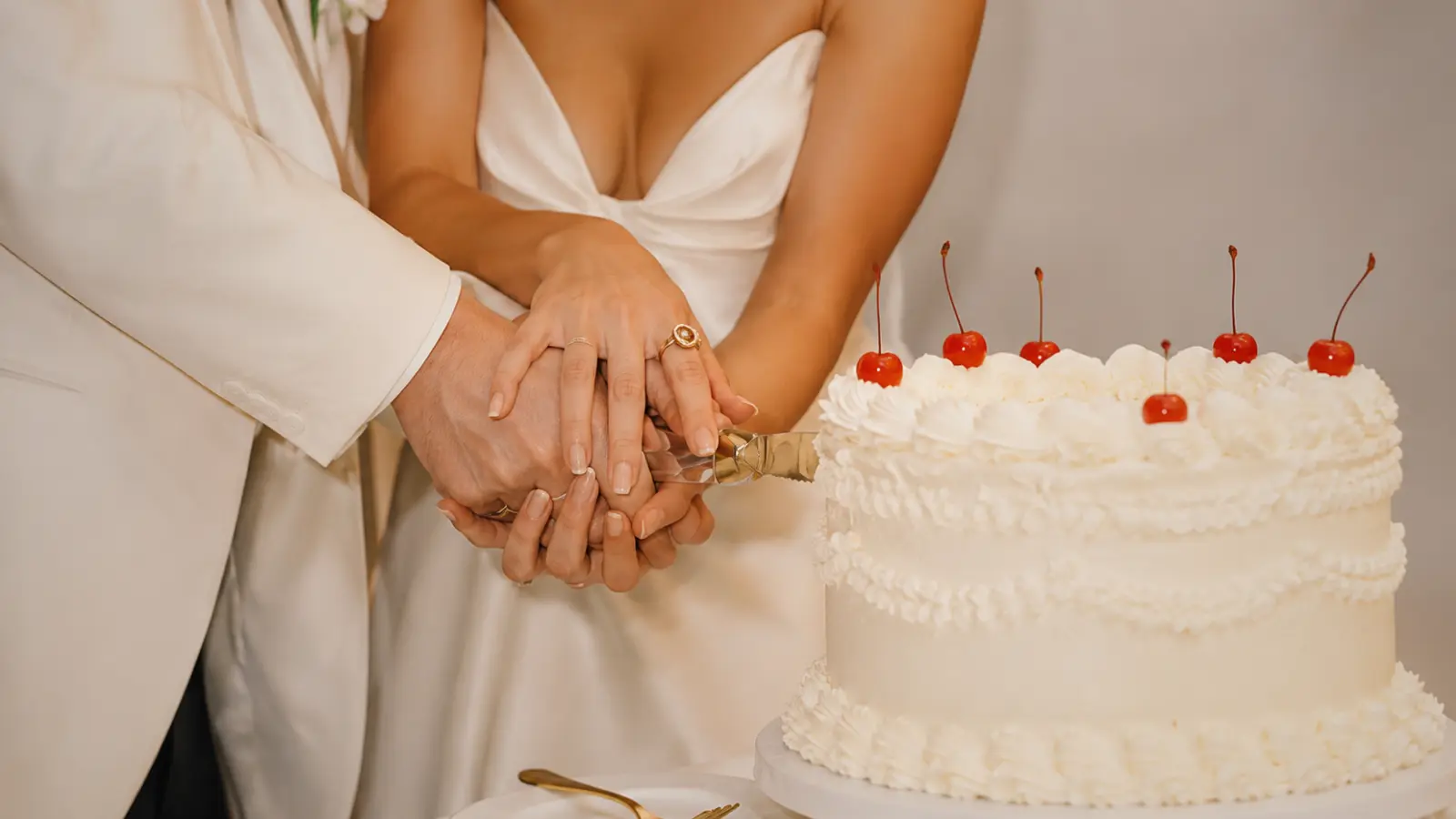 Close-up of bride and groom hands holding a cake knife together while cutting a traditional white wedding cake with cherries.