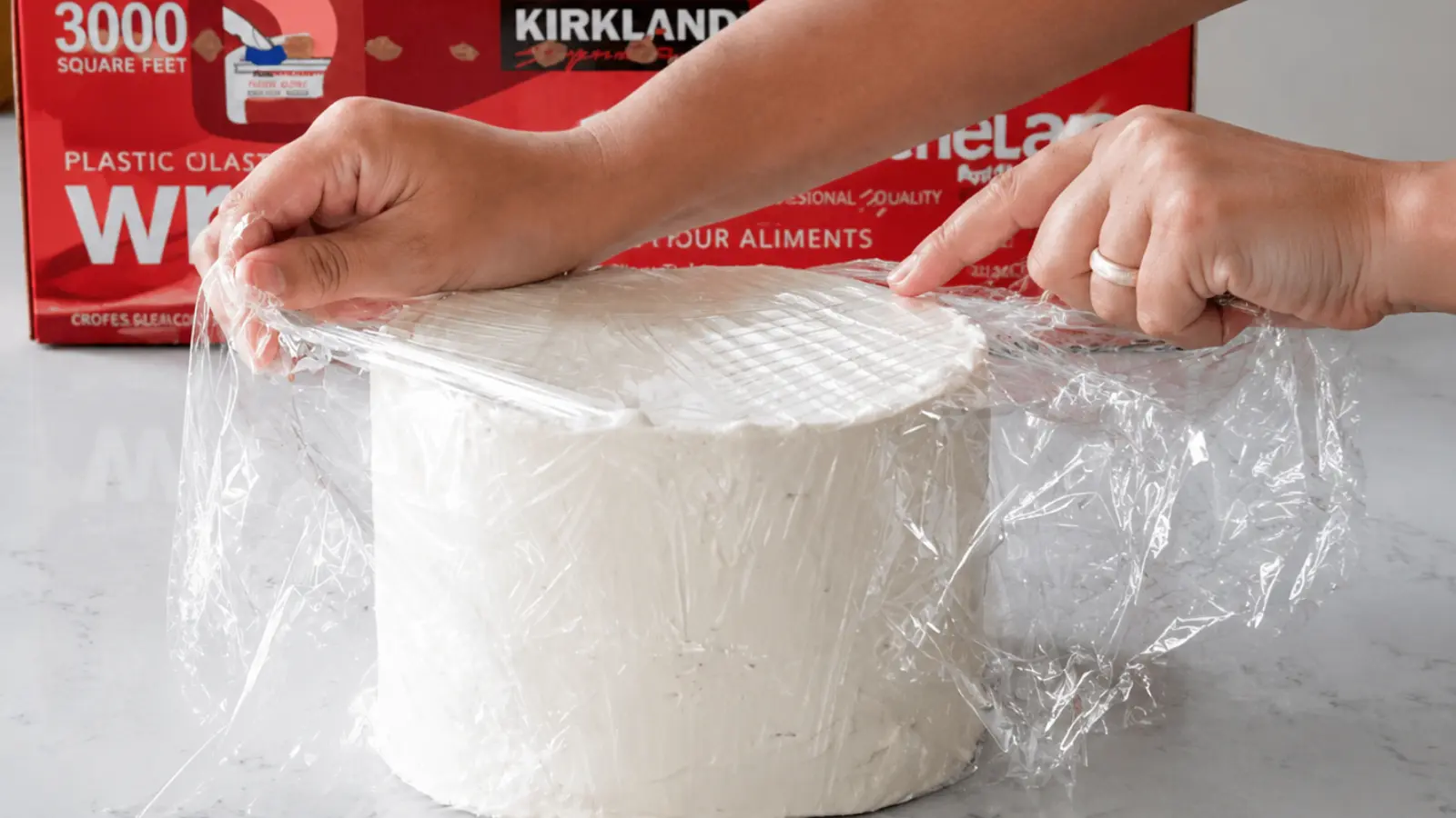 Hands carefully wrapping a wedding cake tier with plastic wrap to protect the frosting and preserve the cake before freezing.