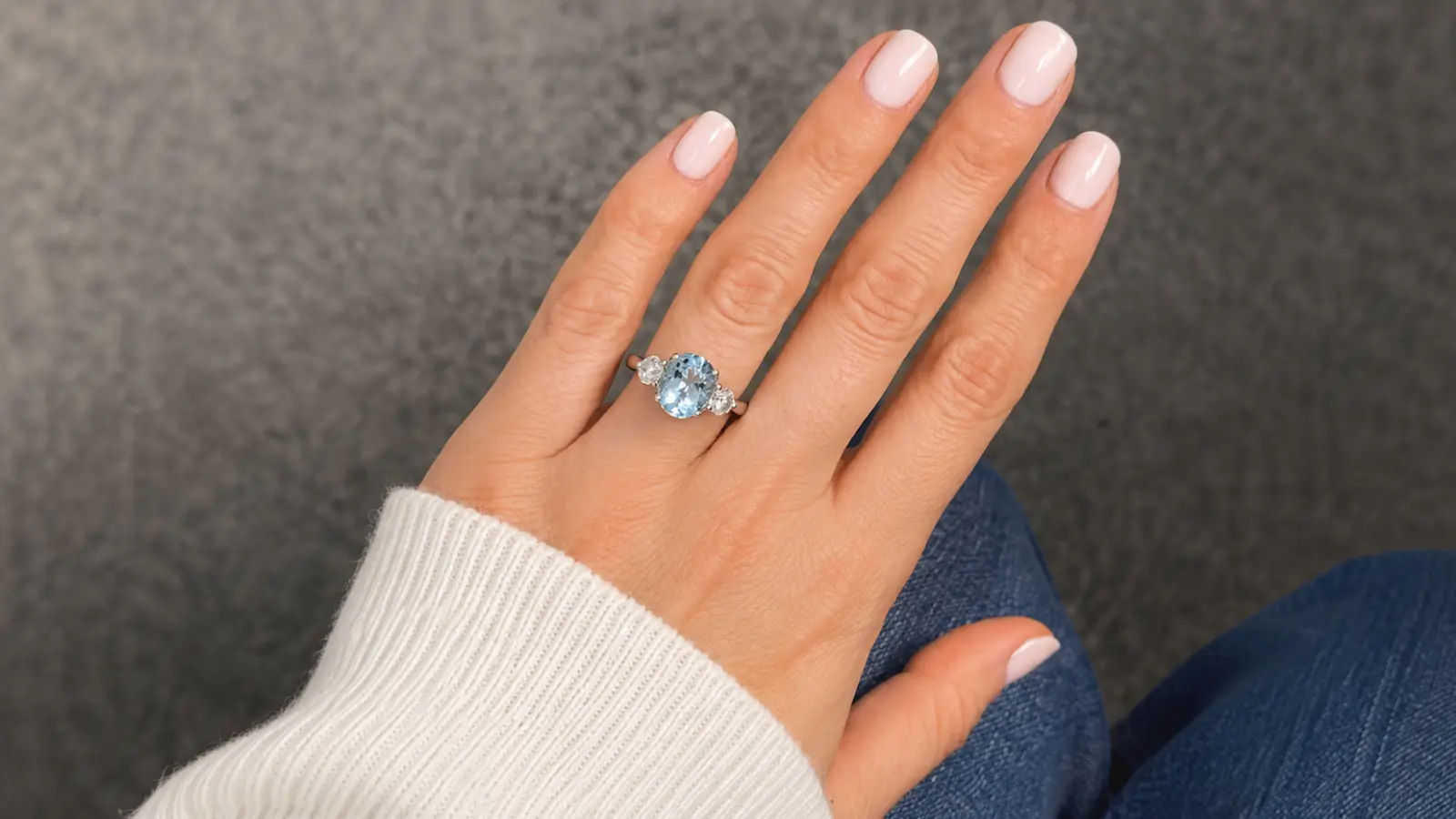 A close-up of a hand wearing an aquamarine gemstone engagement ring with diamond side stones against a casual neutral background