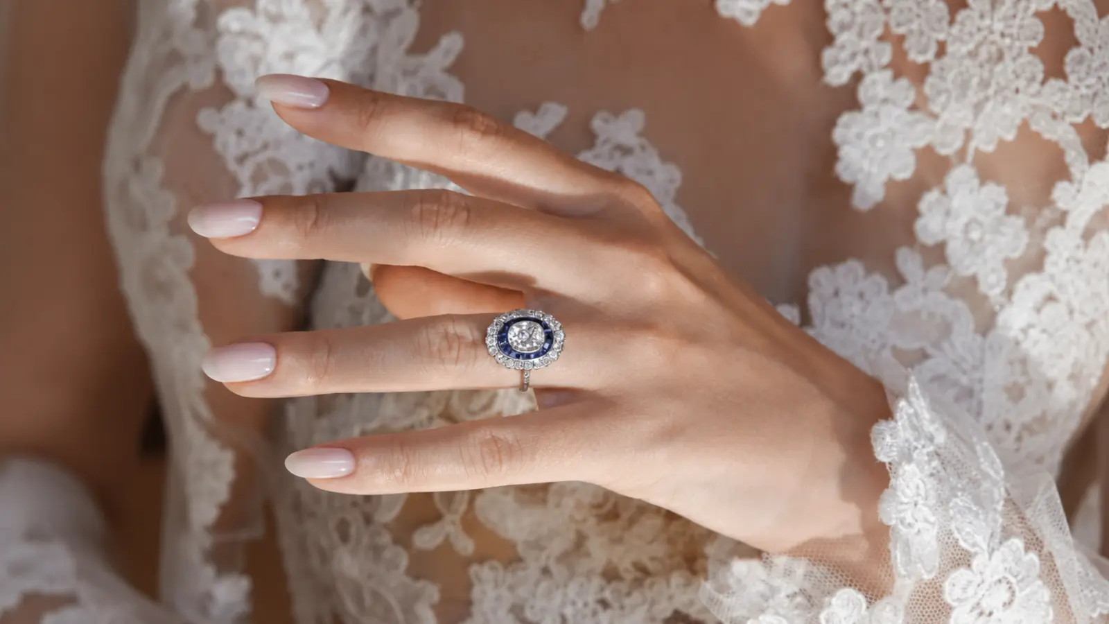 A bride wearing an Art Deco sapphire halo engagement ring with a diamond center and deep blue sapphire accents against lace sleeves