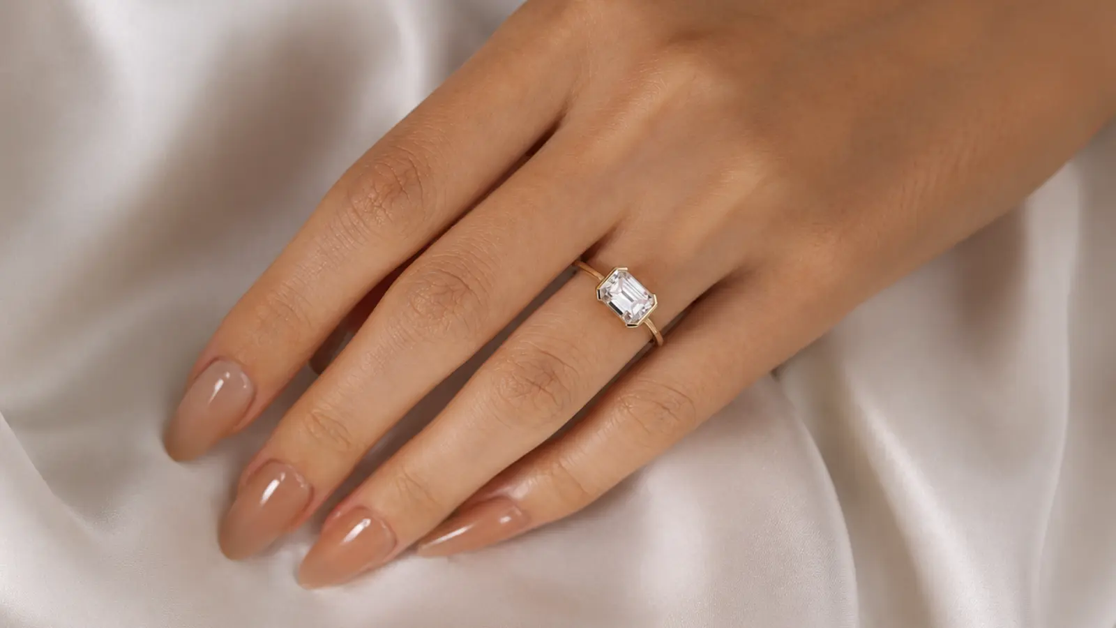 A bezel set emerald cut engagement ring worn on a woman's hand with a minimalist gold band