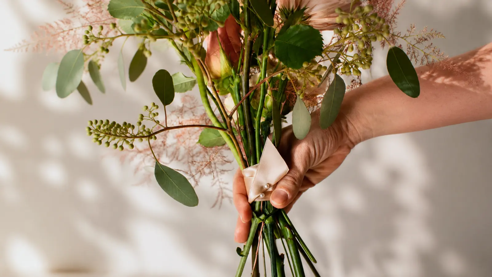 Hand holding the stems of a handmade wedding bouquet secured with ribbon before the final bouquet wrap