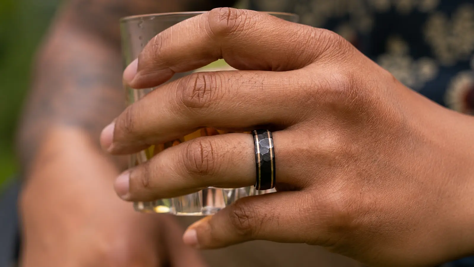 A man wearing a black hammered wedding band with a modern textured finish while holding a glass outdoors