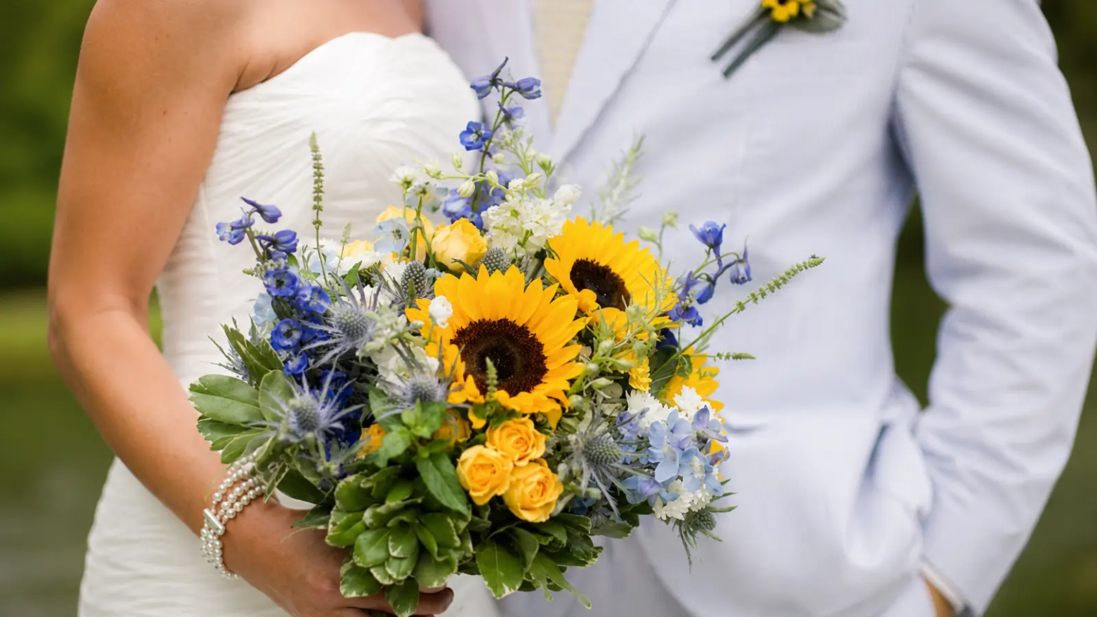 Bride holding a sunflower wedding bouquet with blue delphinium, thistle, yellow roses, and greenery