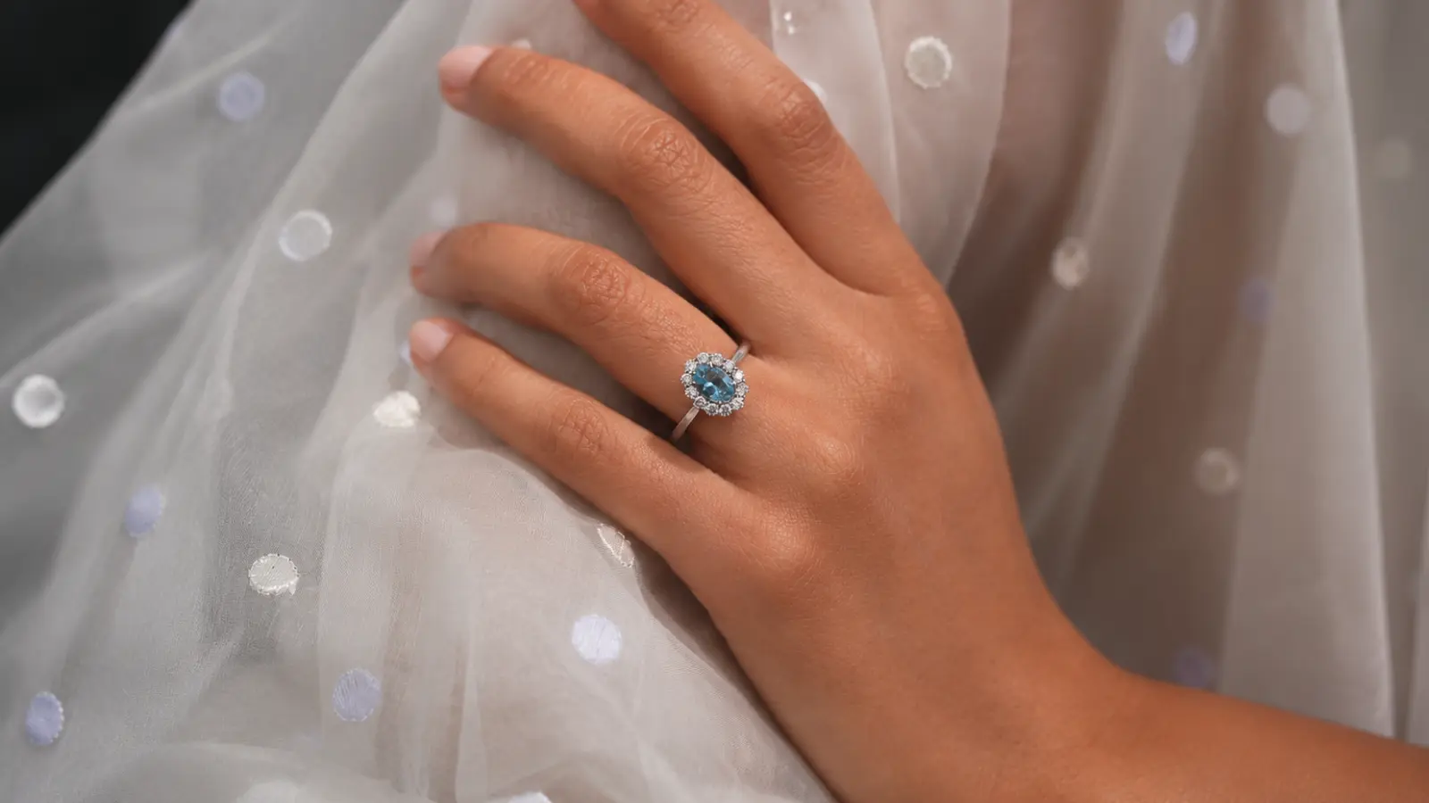 A hand rests on soft fabric while wearing a blue topaz gemstone halo ring surrounded by sparkling accent stones on a white metal band