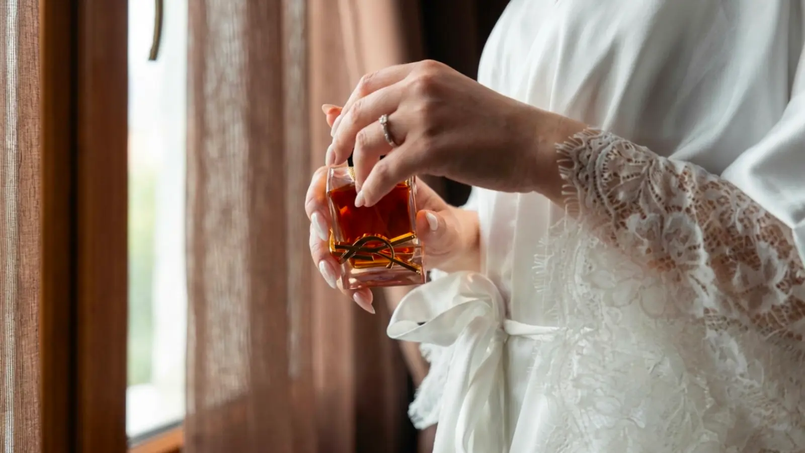 A bride holding an elegant wedding perfume near a window while preparing for the ceremony, choosing a signature bridal fragrance that reflects her personality and complements the wedding atmosphere