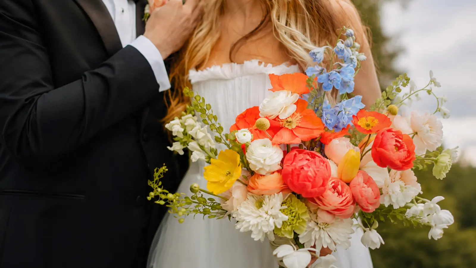 Bride holding a bright multicolor wedding bouquet with coral peonies, poppies and blue delphiniums