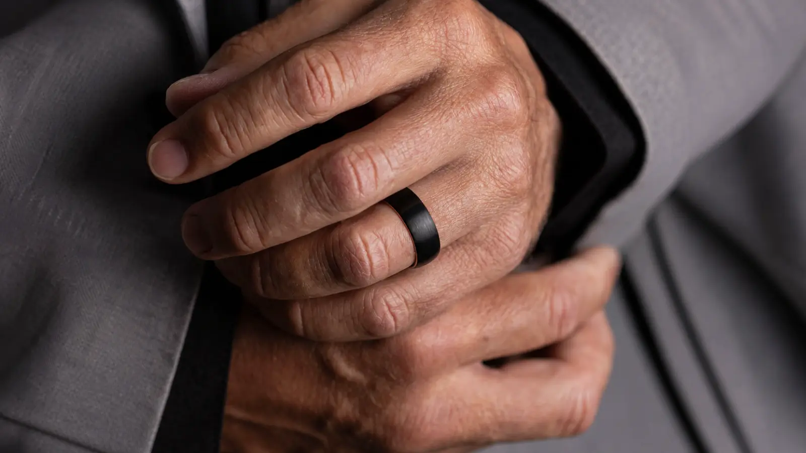 A close-up of a brushed black men's wedding band on a hand wearing a tailored gray suit jacket
