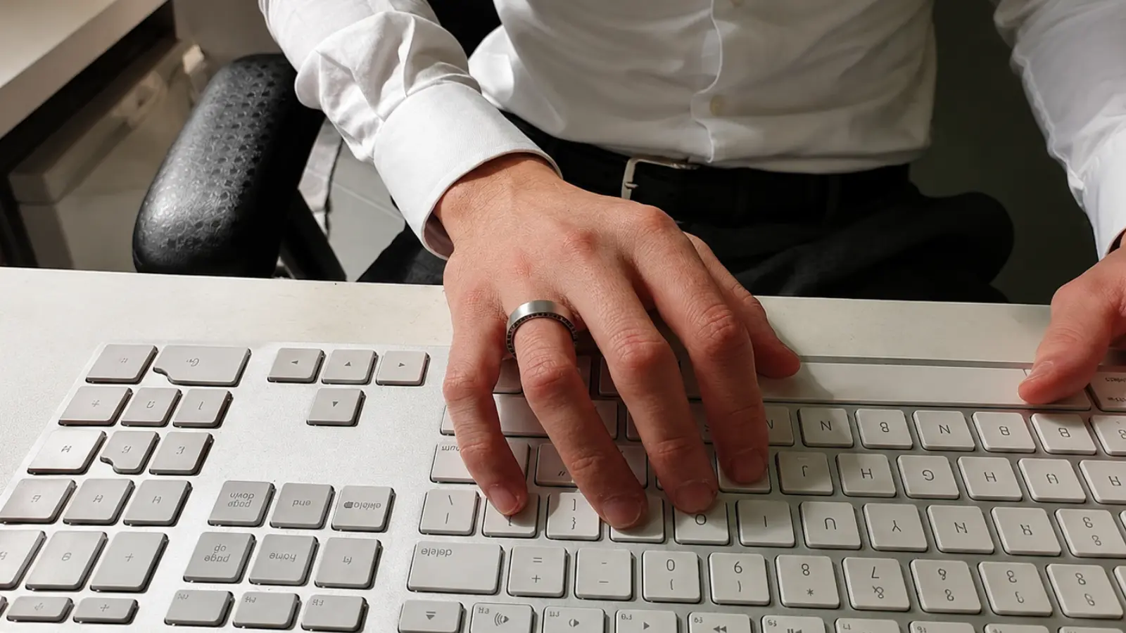 Close-up of a man's hand wearing a brushed titanium men's wedding ring while typing on a keyboard, highlighting a durable everyday wedding band for work environments
