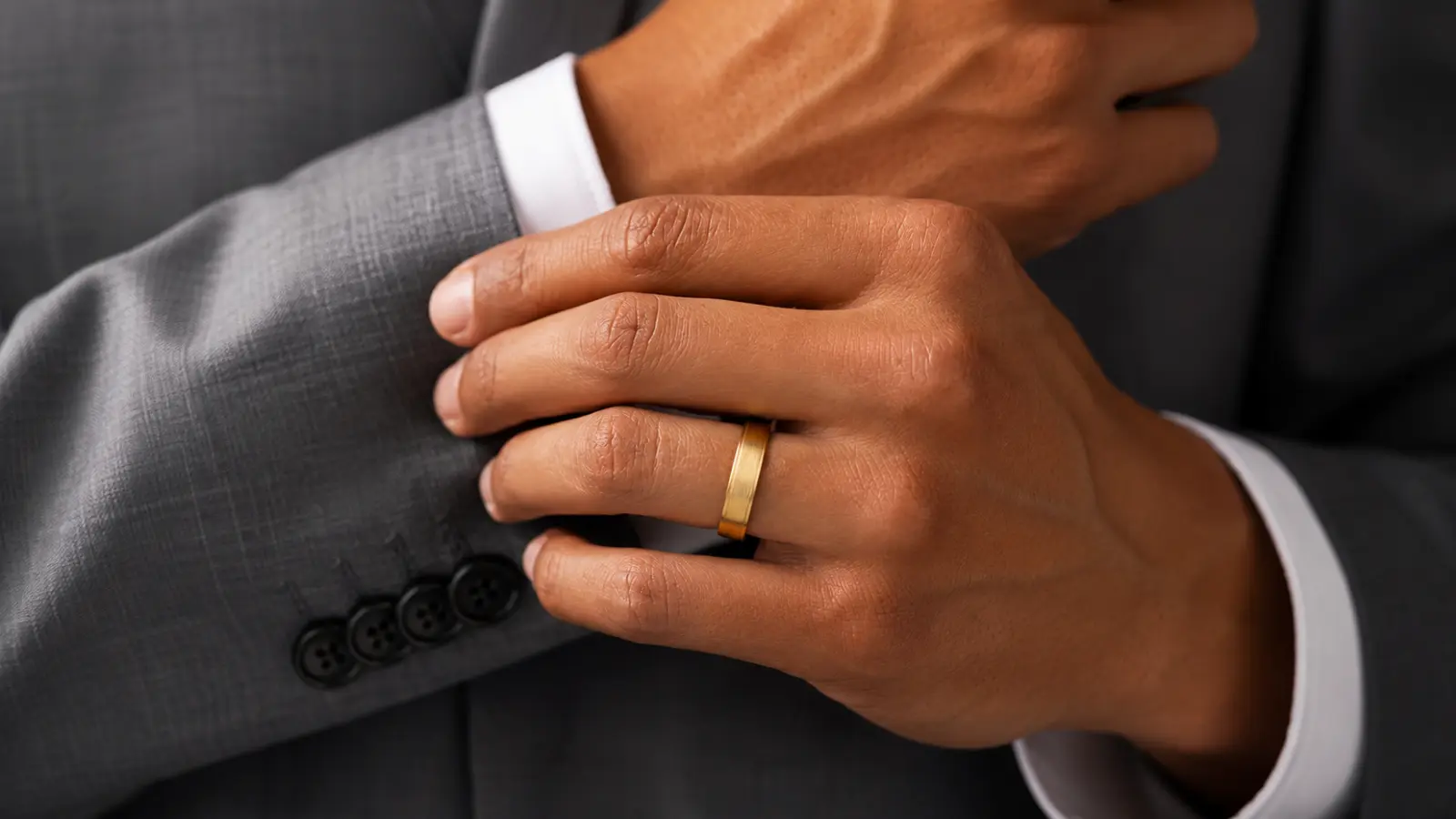 Groom adjusting the sleeve of a gray suit while wearing a brushed yellow gold men's wedding band, showcasing a classic masculine wedding ring with a refined finish