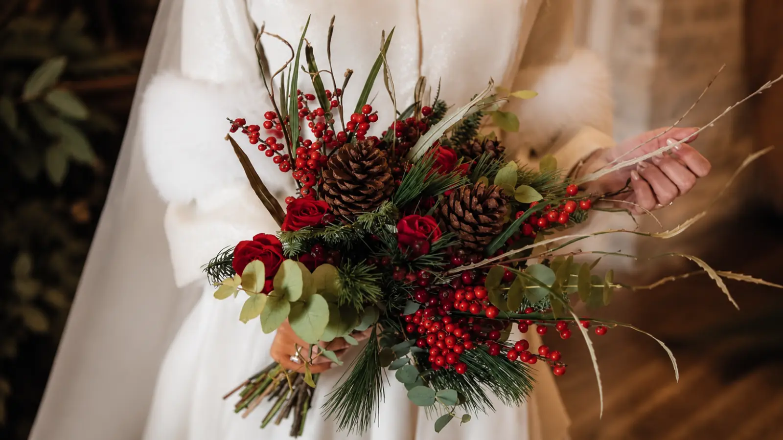 Bride holding a Christmas winter wedding bouquet with pinecones, red berries, evergreen branches and red roses