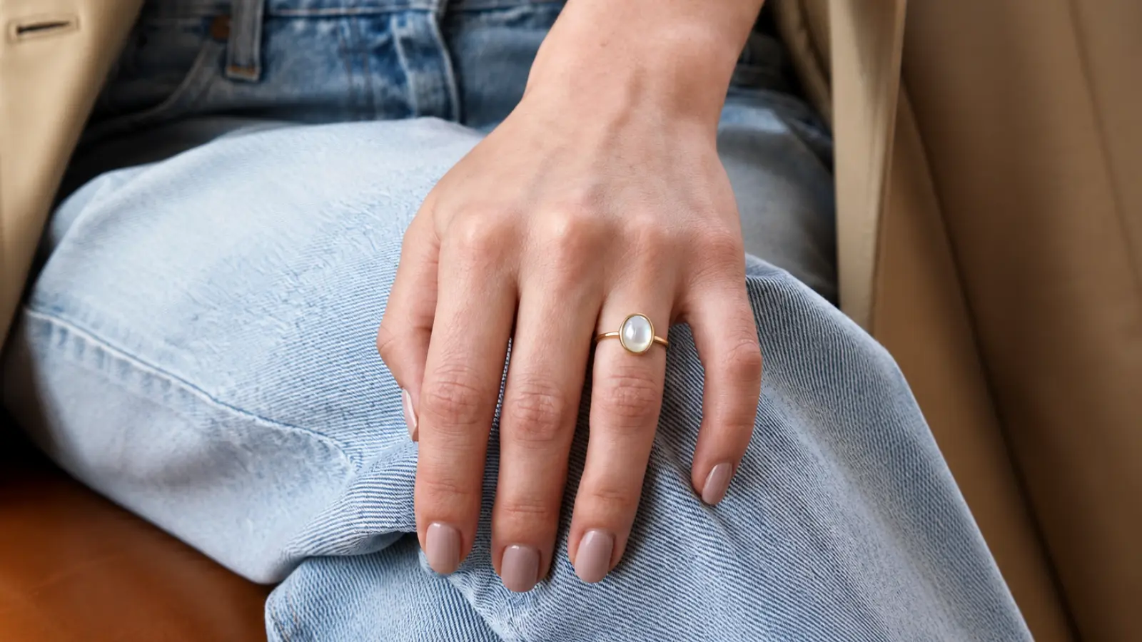 A hand resting on denim wears a classic opal solitaire ring with a luminous oval opal set in a simple yellow gold bezel setting