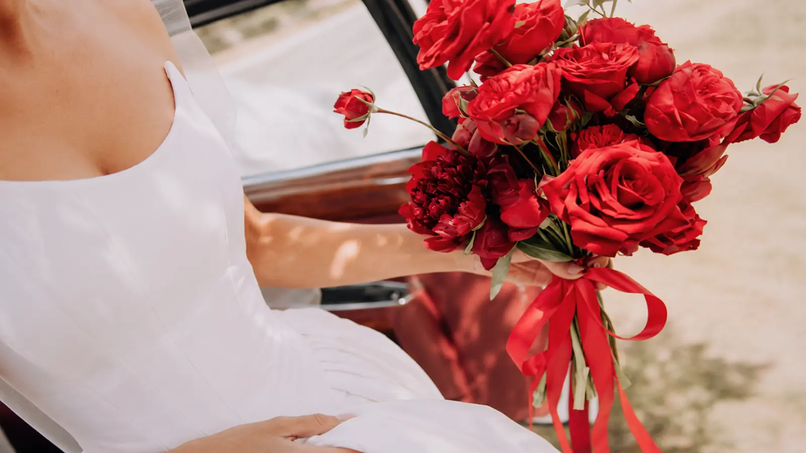 Bride holding a timeless red rose wedding bouquet tied with a long satin ribbon beside a vintage car