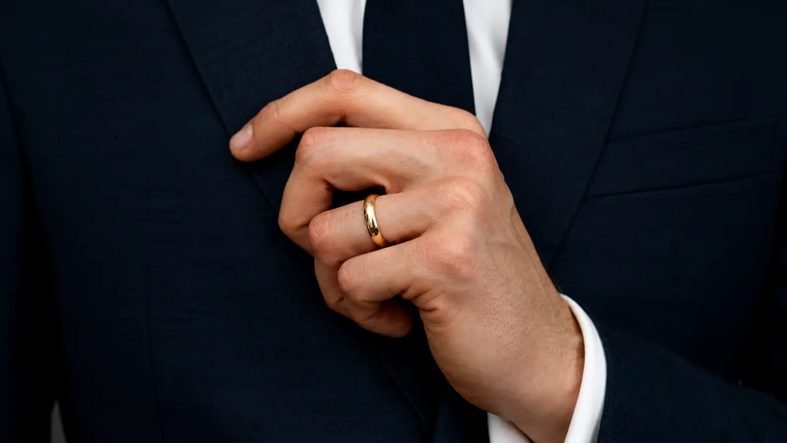 A man wearing a classic polished yellow gold wedding band while adjusting the lapel of a navy blue suit Wedding Ring Styles