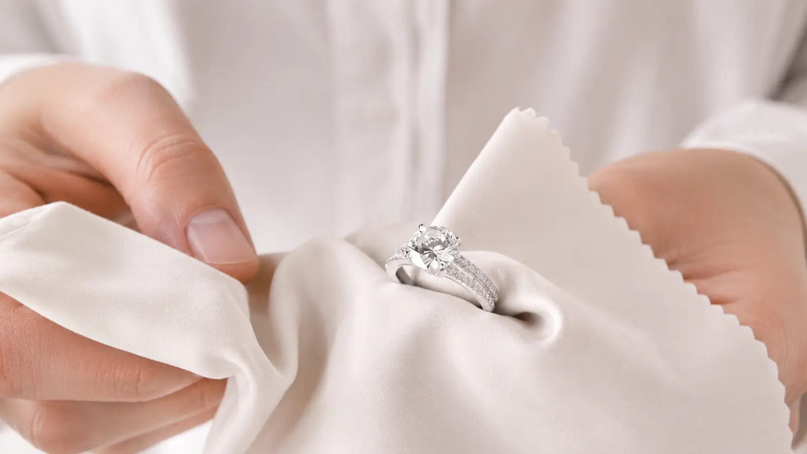 Hands carefully clean a silver engagement ring with a soft polishing cloth, highlighting a simple jewelry care routine that helps preserve its polished appearance