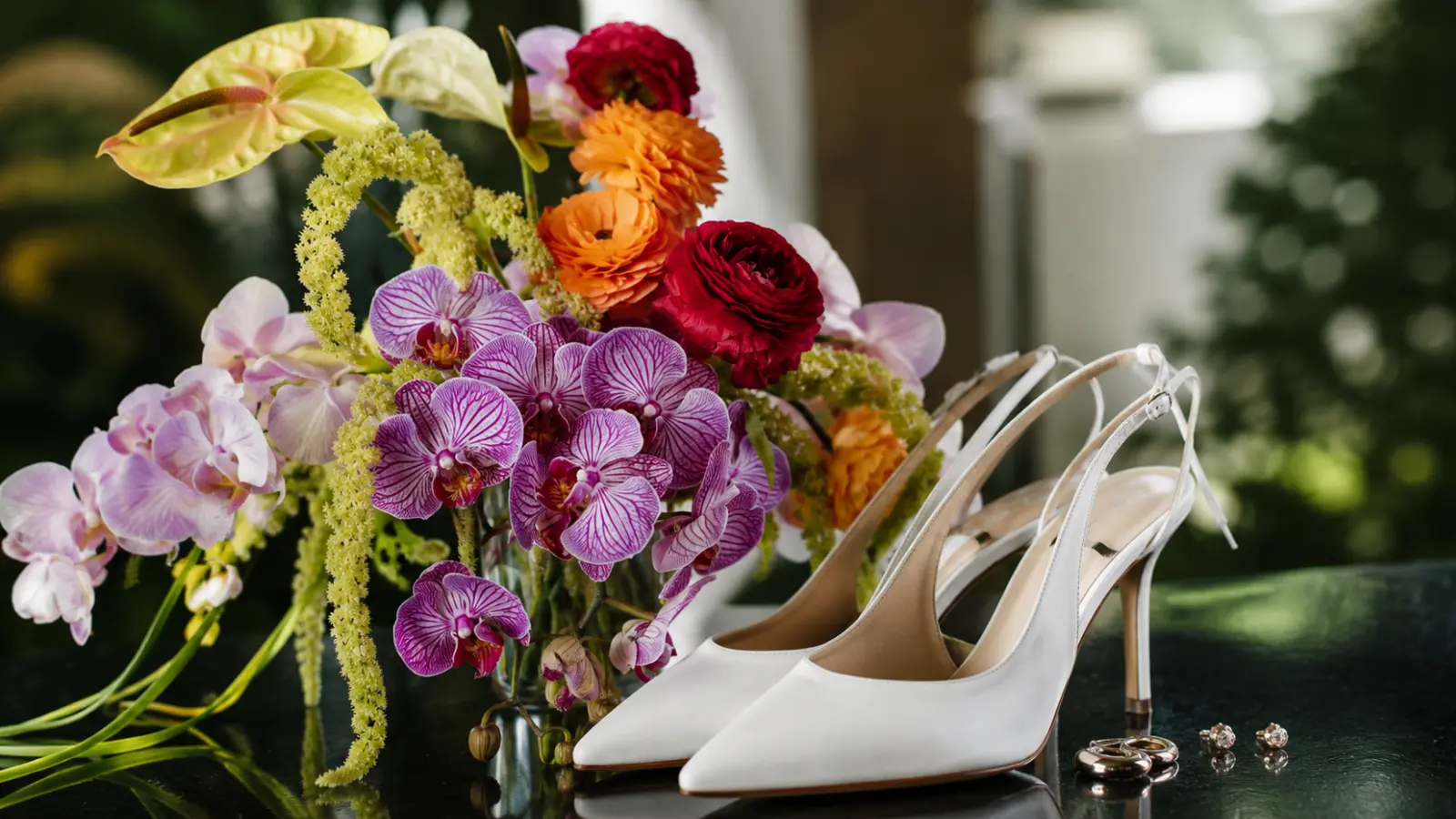 Colorful orchid and anthurium wedding bouquet displayed beside white bridal heels, wedding rings, and earrings