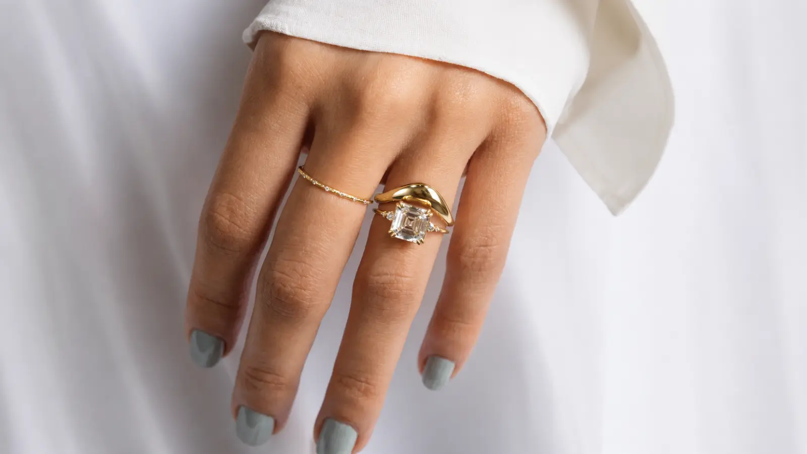 A hand displaying a curved wedding band stacked with an emerald-cut engagement ring and complementary yellow gold bands Curved Wedding Bands