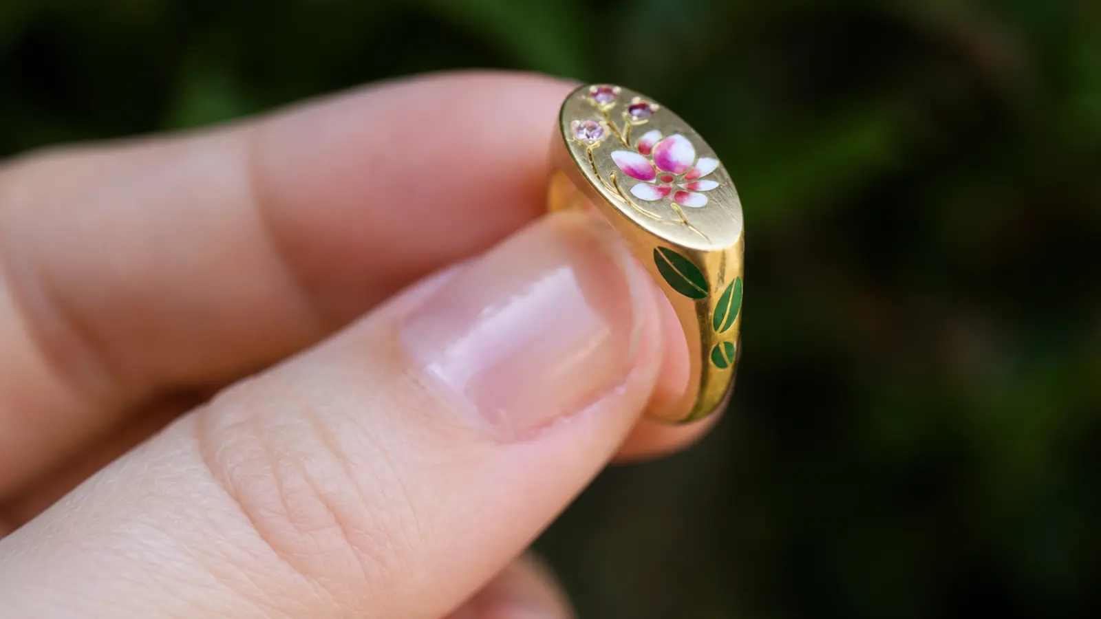 A custom gold ring with floral enamel details held between fingers to showcase its handcrafted decorative design
