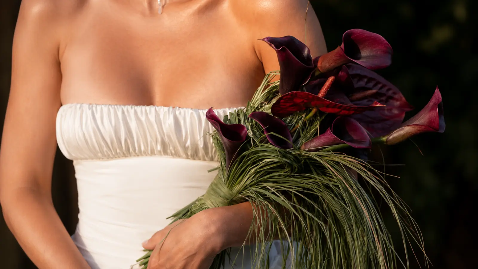 Bride holding a dark calla lily wedding bouquets with burgundy flowers, tropical greenery, and a modern strapless dress