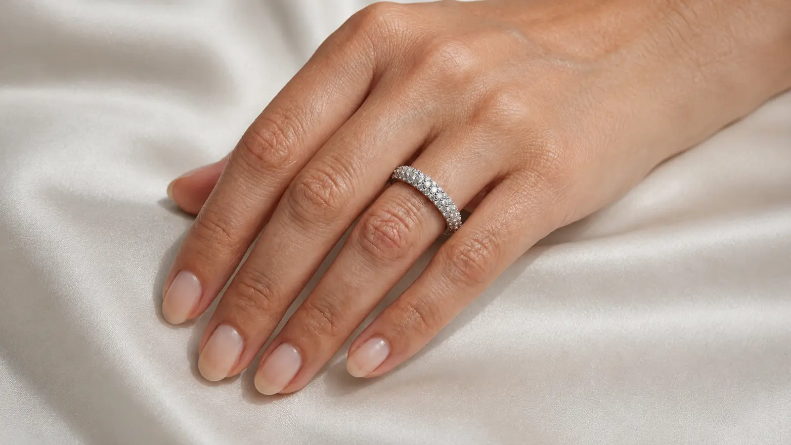 A hand wearing a diamond eternity wedding band with round diamonds encircling the band on a soft white satin background