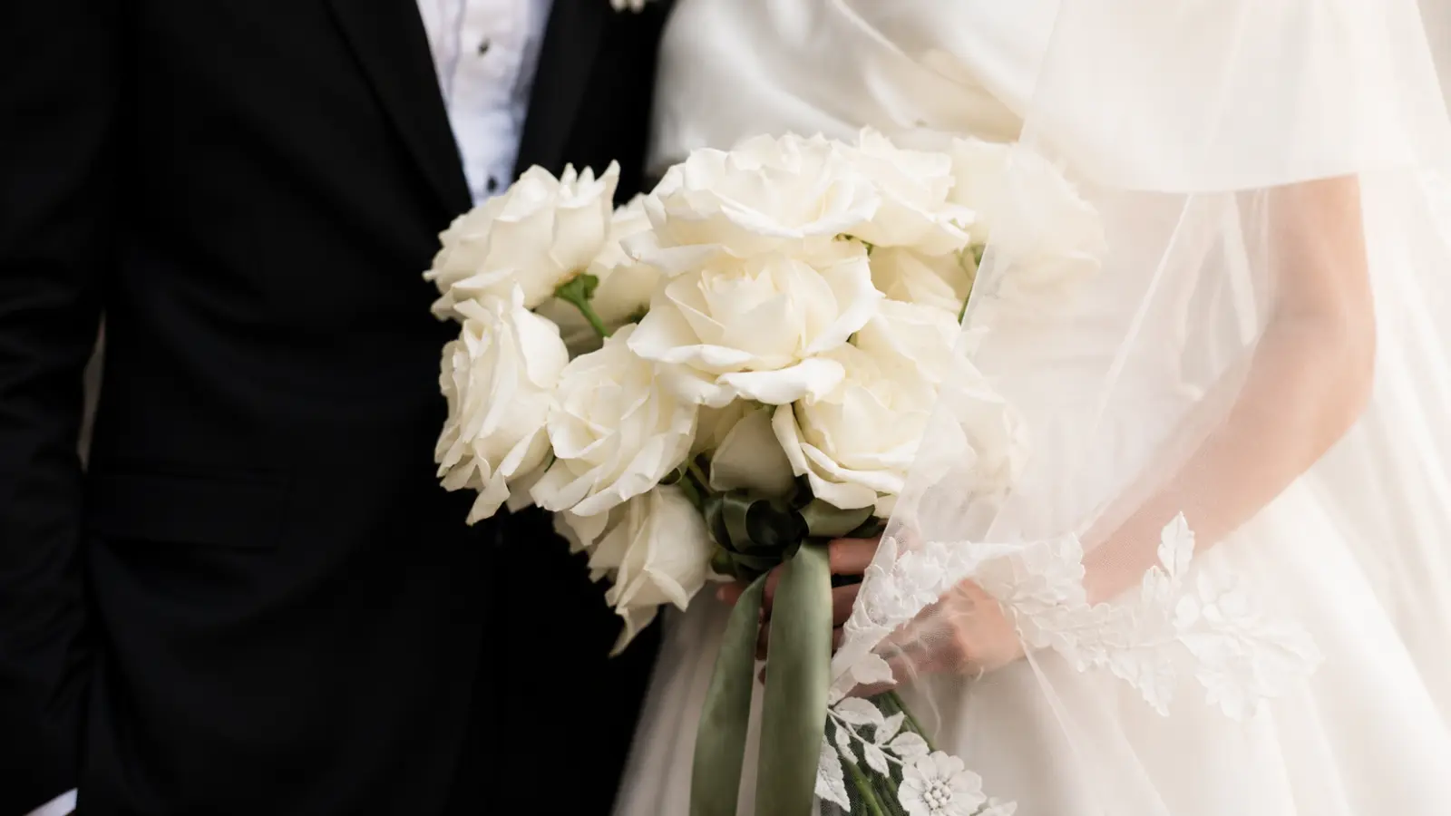 Bride and groom holding an elegant white rose wedding bouquet finished with a soft green ribbon during their wedding