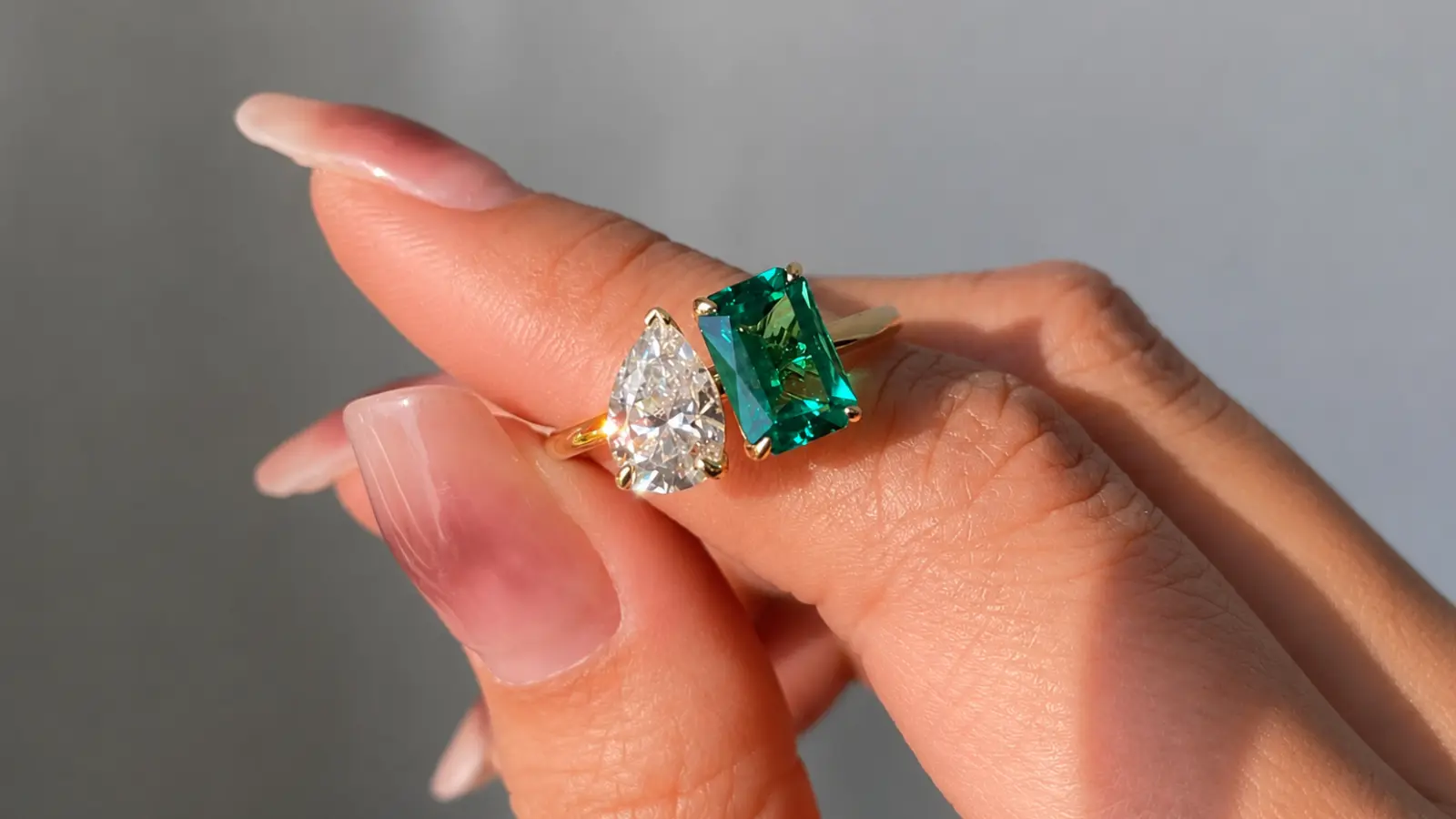 A close-up of an emerald and diamond two-stone engagement ring featuring a rectangular emerald and pear-shaped diamond in yellow gold