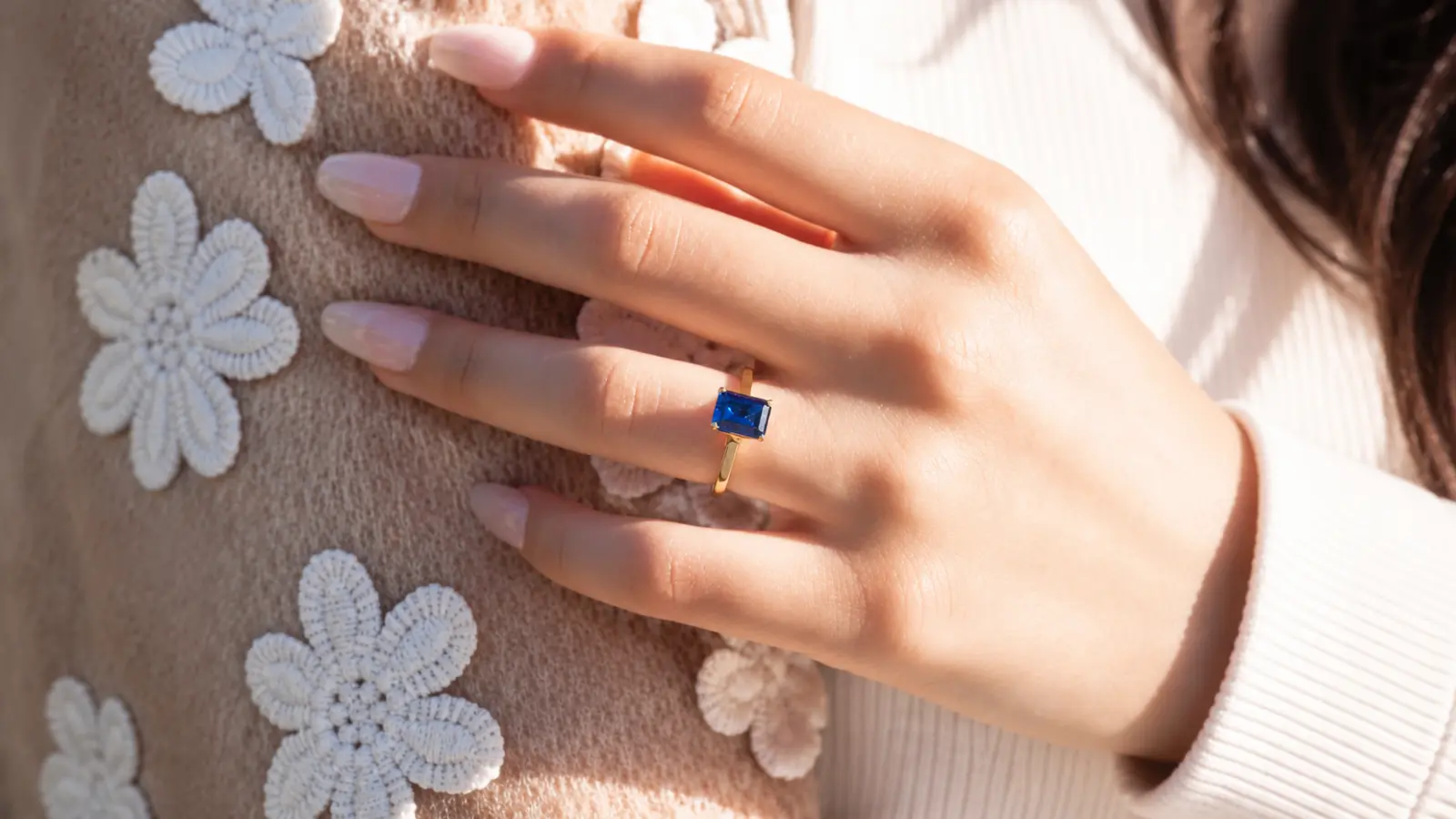 A woman wears an emerald cut sapphire solitaire ring with a vivid blue gemstone set on a sleek yellow gold band against a floral sweater