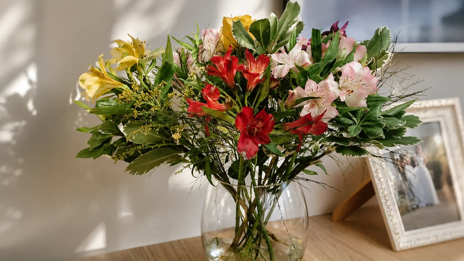 Finished DIY wedding bouquet displayed in a glass vase with colorful fresh flowers and greenery beside a framed wedding photo