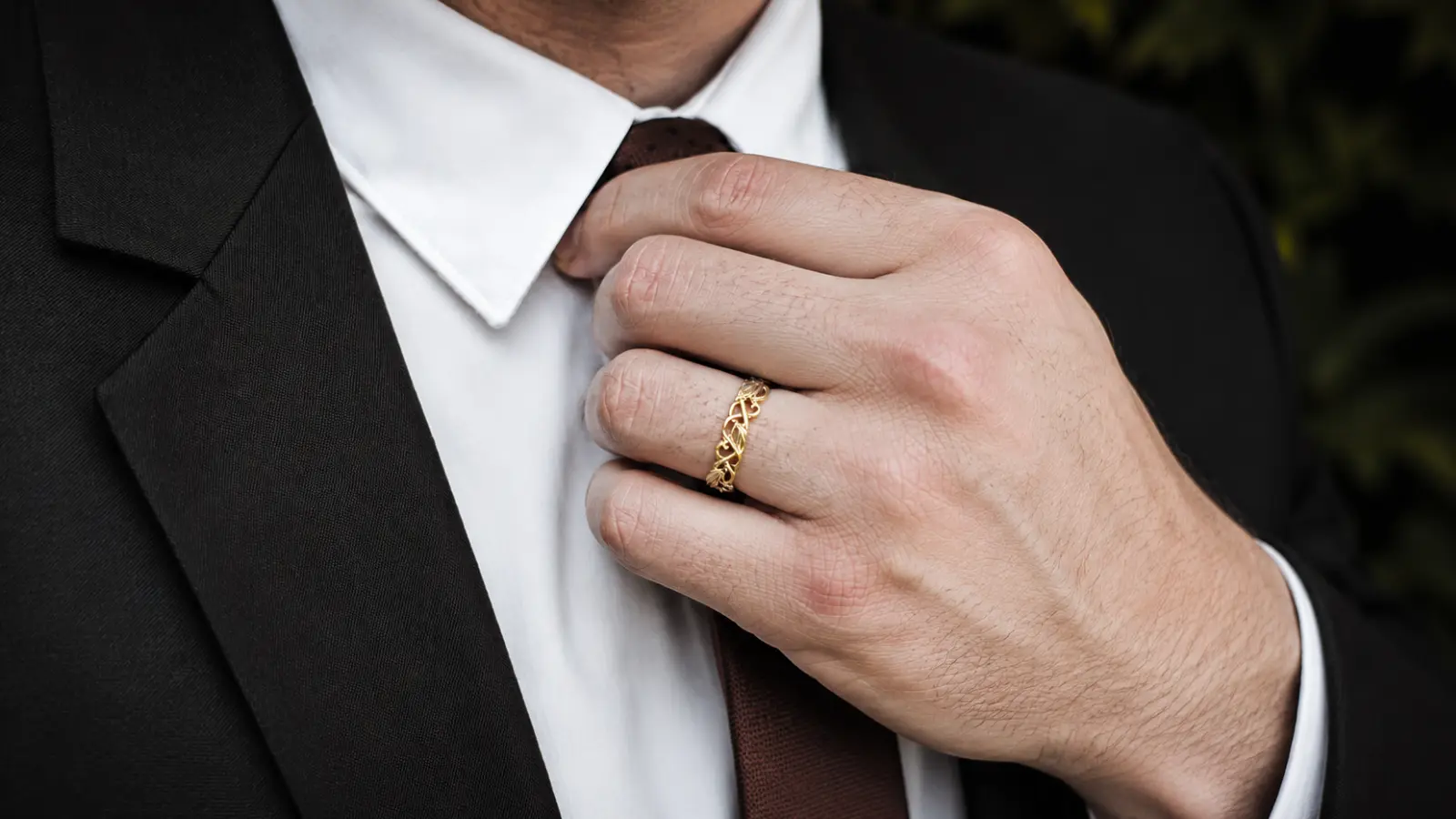A man adjusting his tie while wearing a yellow gold floral vine wedding band with intricate botanical detailing