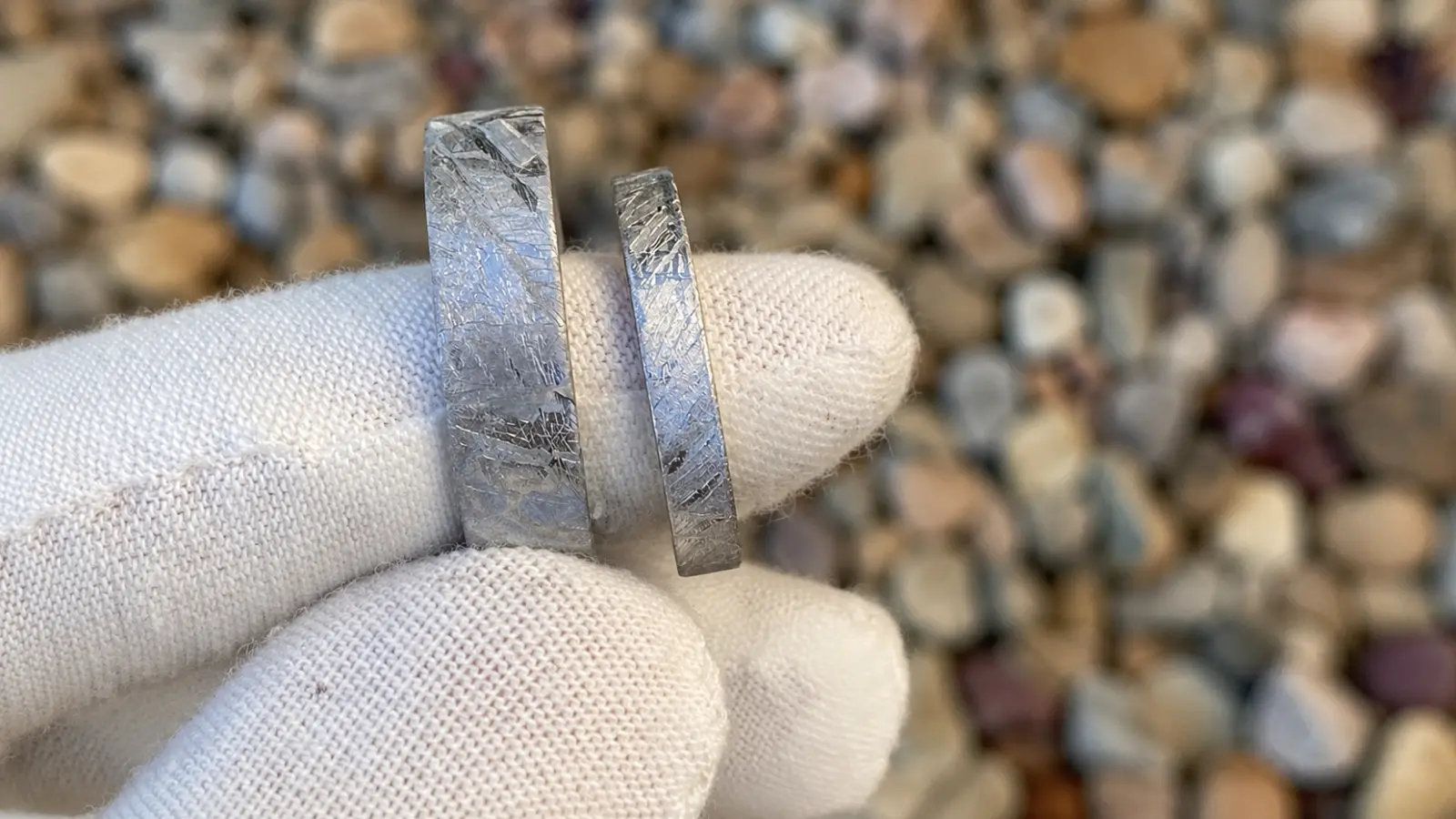 A gloved hand holds two genuine meteorite wedding rings, clearly displaying the distinctive crystalline Widmanstätten pattern found in authentic meteorite material