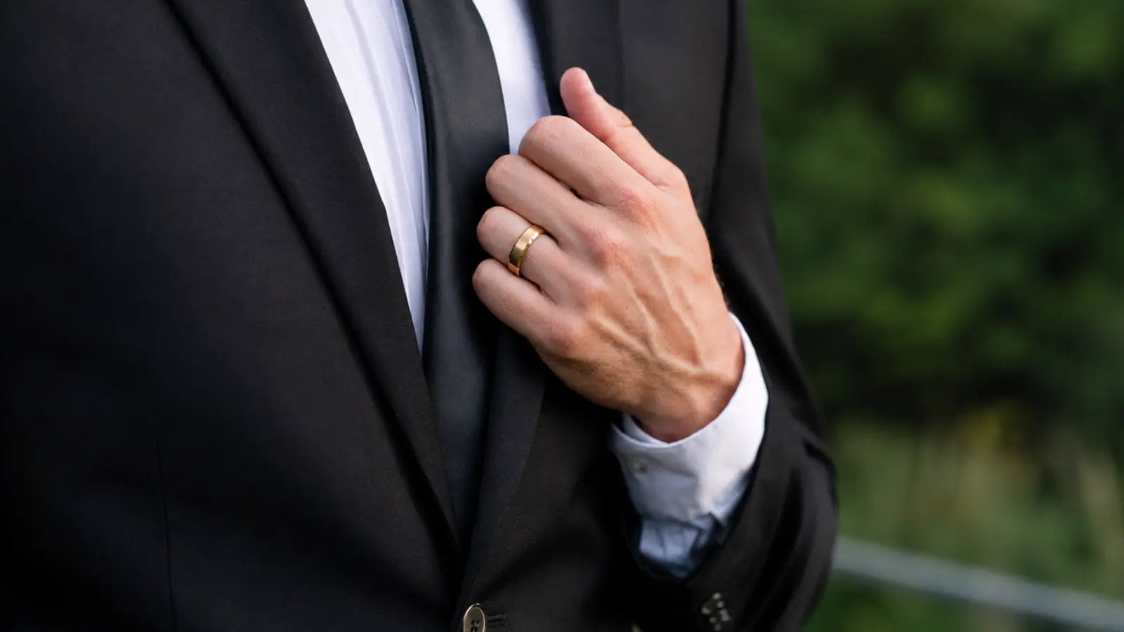 A groom wearing a black tuxedo gently adjusts his lapel while displaying a polished yellow gold wedding ring, highlighting the classic style and versatility of a traditional men's wedding band