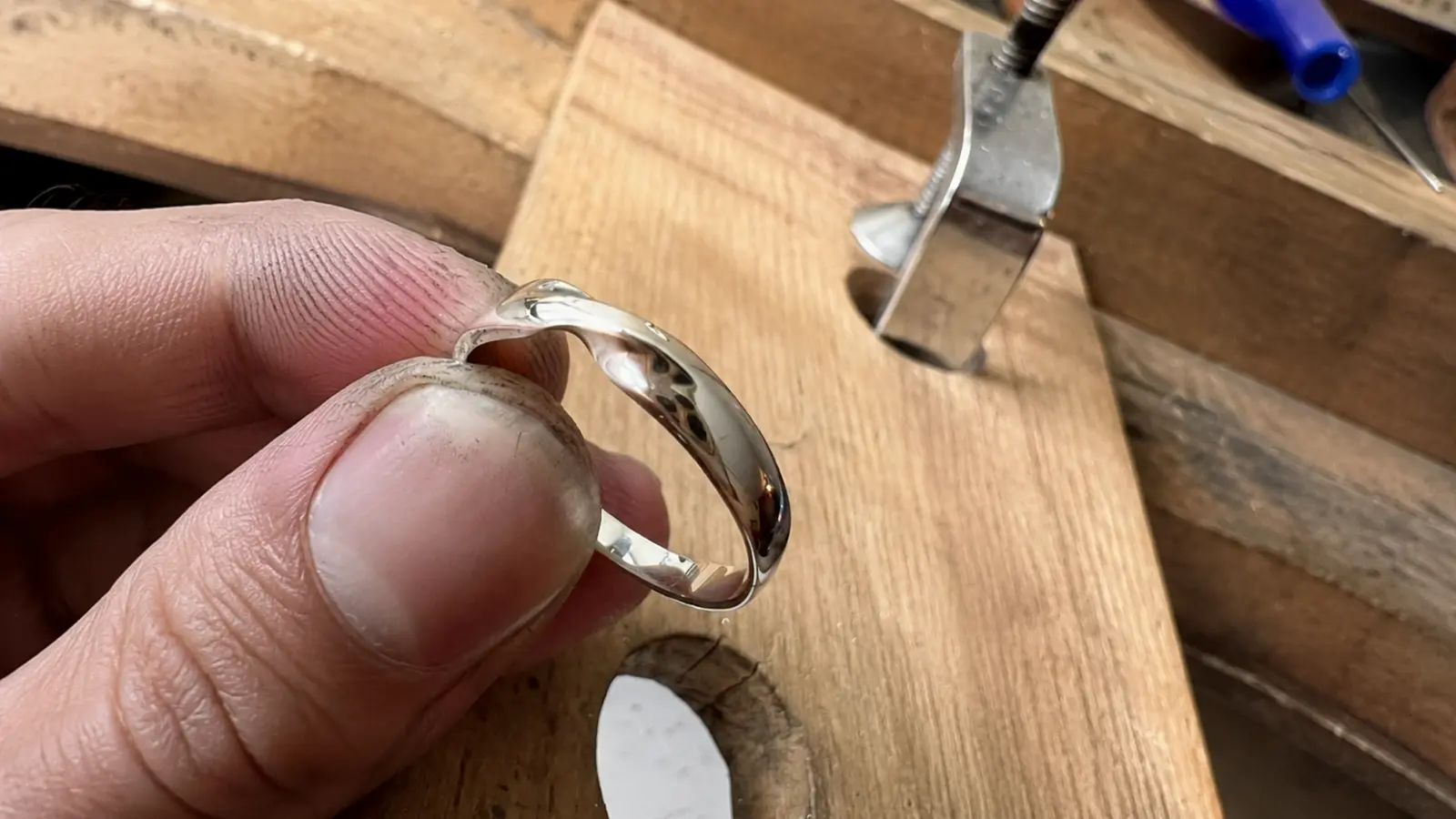 A jeweler holds a handcrafted white gold wedding ring above a wooden workbench, showcasing the detailed craftsmanship, smooth polish and precision involved in creating a high-quality wedding band
