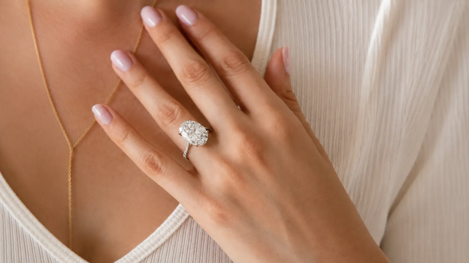 A close-up of a hand wearing a large oval moissanite engagement ring with a delicate pavé band against a neutral outfit