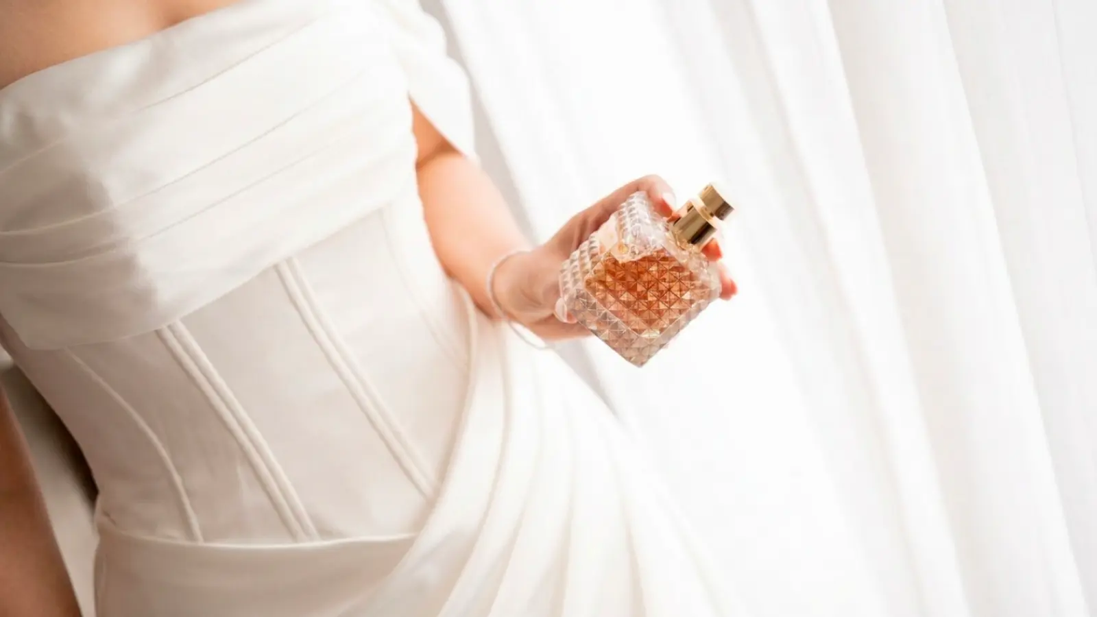 A luxury bridal perfume held beside an elegant wedding dress, showing how the perfect fragrance becomes an essential finishing touch that enhances the bride's style and wedding day experience