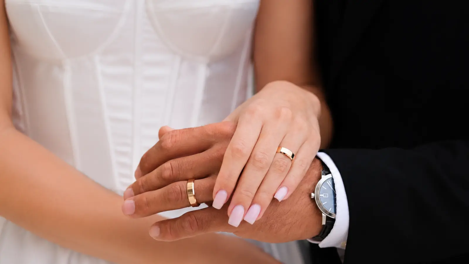 A bride rests her hand on the groom's while both wear matching gold wedding bands, demonstrating a traditional coordinated ring style suitable for modern and classic weddings
