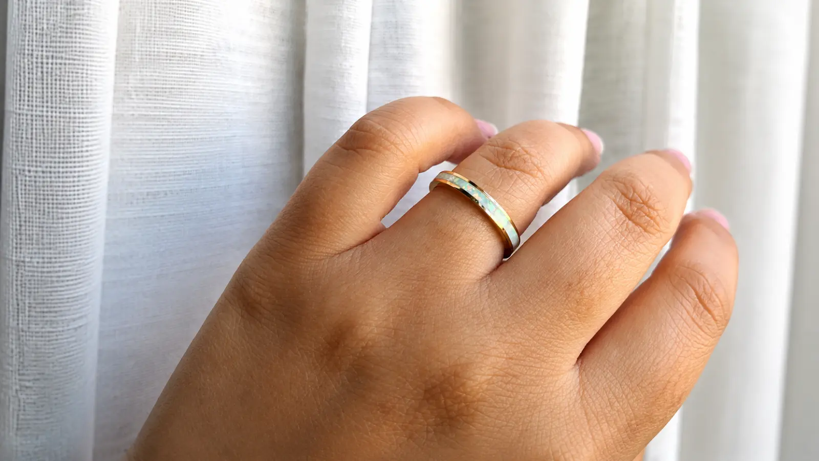A hand wearing a gold wedding band with a luminous opal inlay that displays subtle flashes of blue and green along the band
