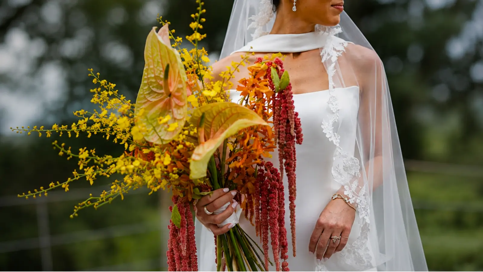 Bride wearing a veil and holding a cascading orange and yellow wedding bouquet with anthuriums, orchids, and red amaranthus