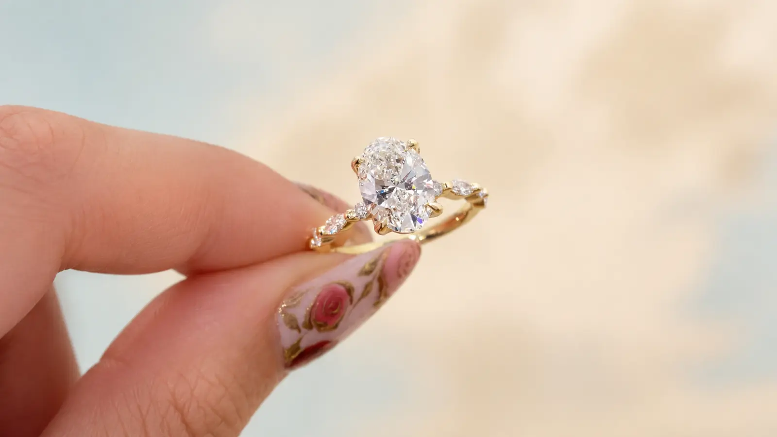 A hand holds an oval moissanite engagement ring with a yellow gold band and marquise side stones in a close-up view